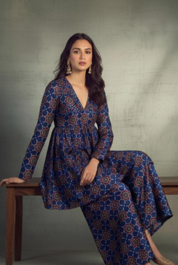 Woman wearing a blue patterned dress sitting on a wooden stool against a gray background