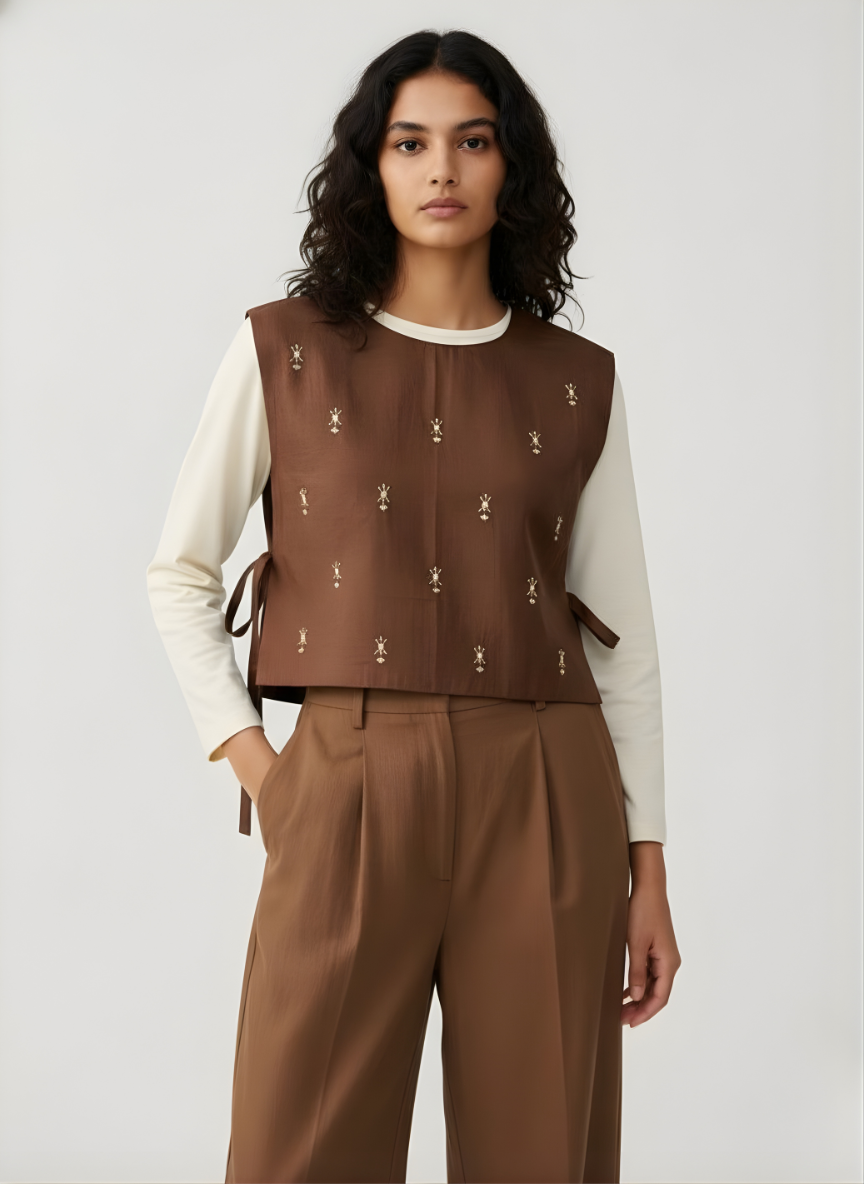 Woman wearing a brown patterned vest over a white long-sleeve shirt with brown pants 