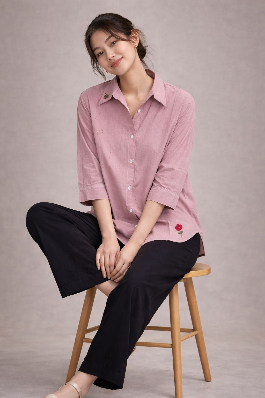 Woman sitting on a stool wearing a pink shirt and black pants against a plain background