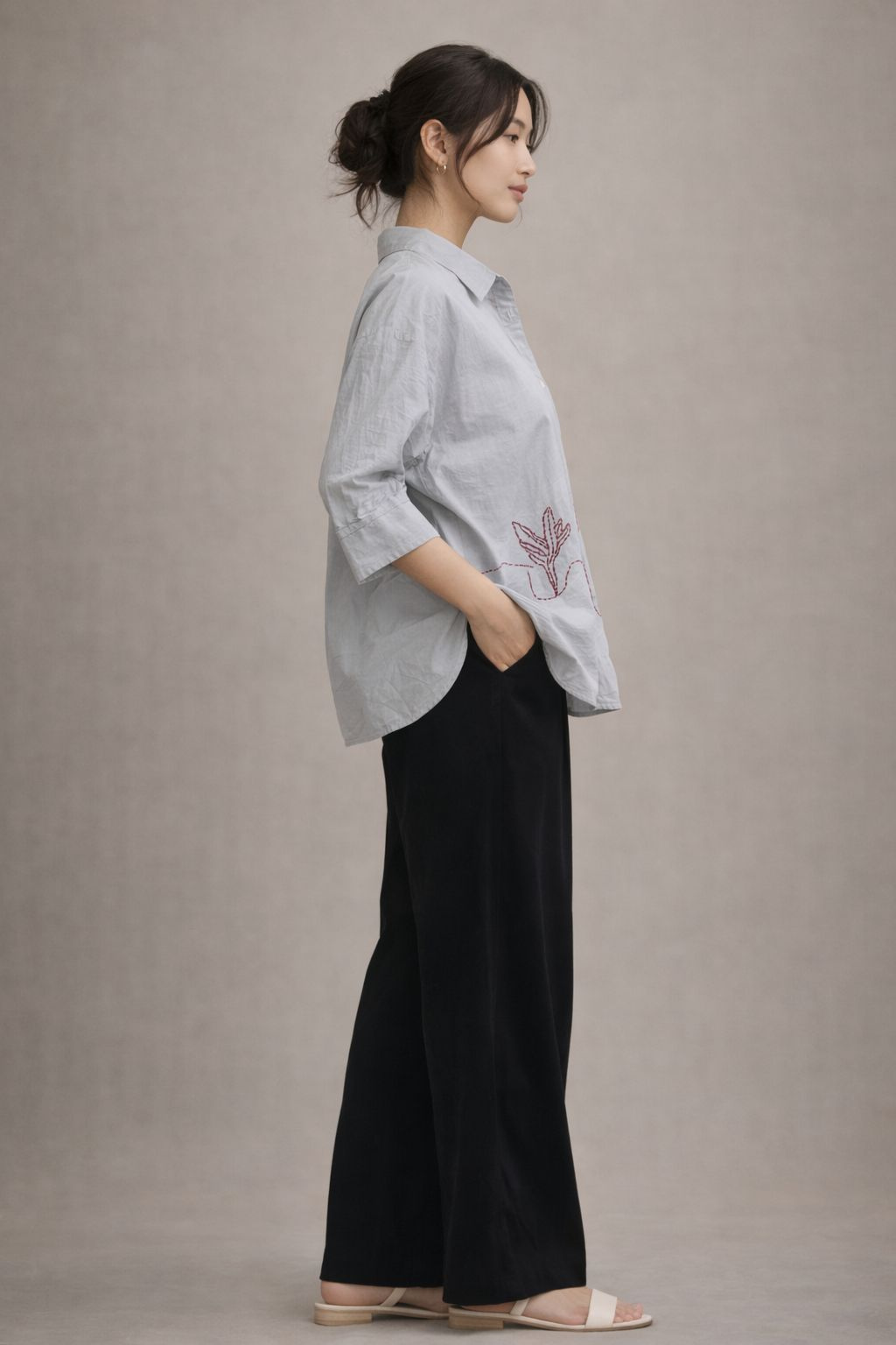 Woman wearing a light gray shirt with floral embroidery and black pants against a plain background