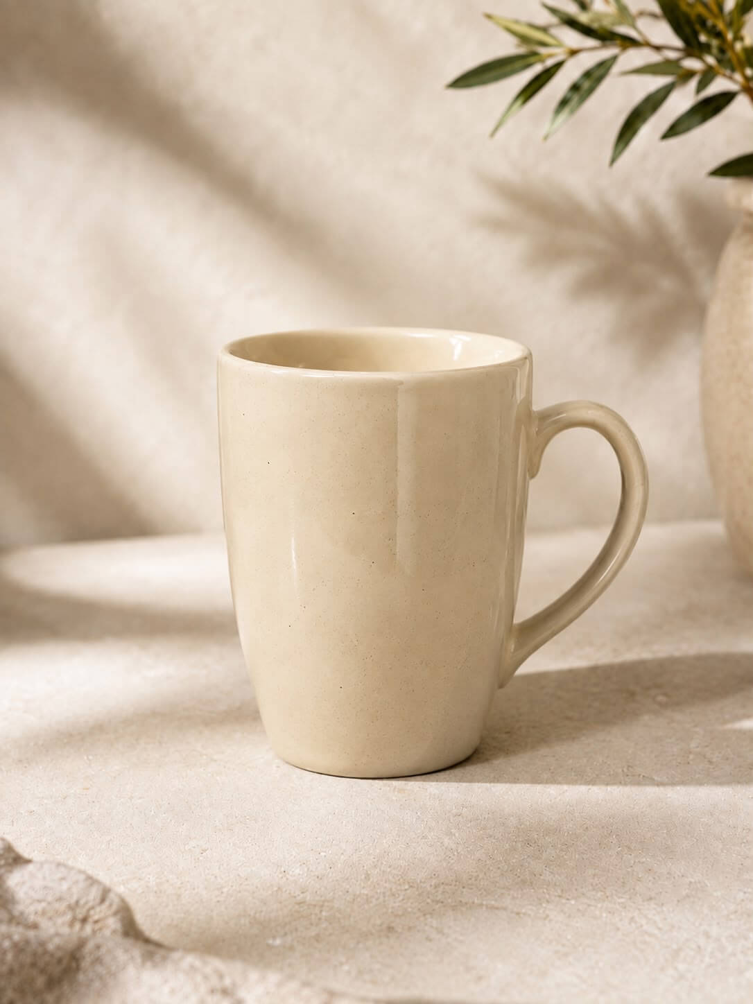 A minimalist ceramic mug in a warm cream or off-white color, styled on a textured beige surface with soft leaf shadows and a plant in the background.