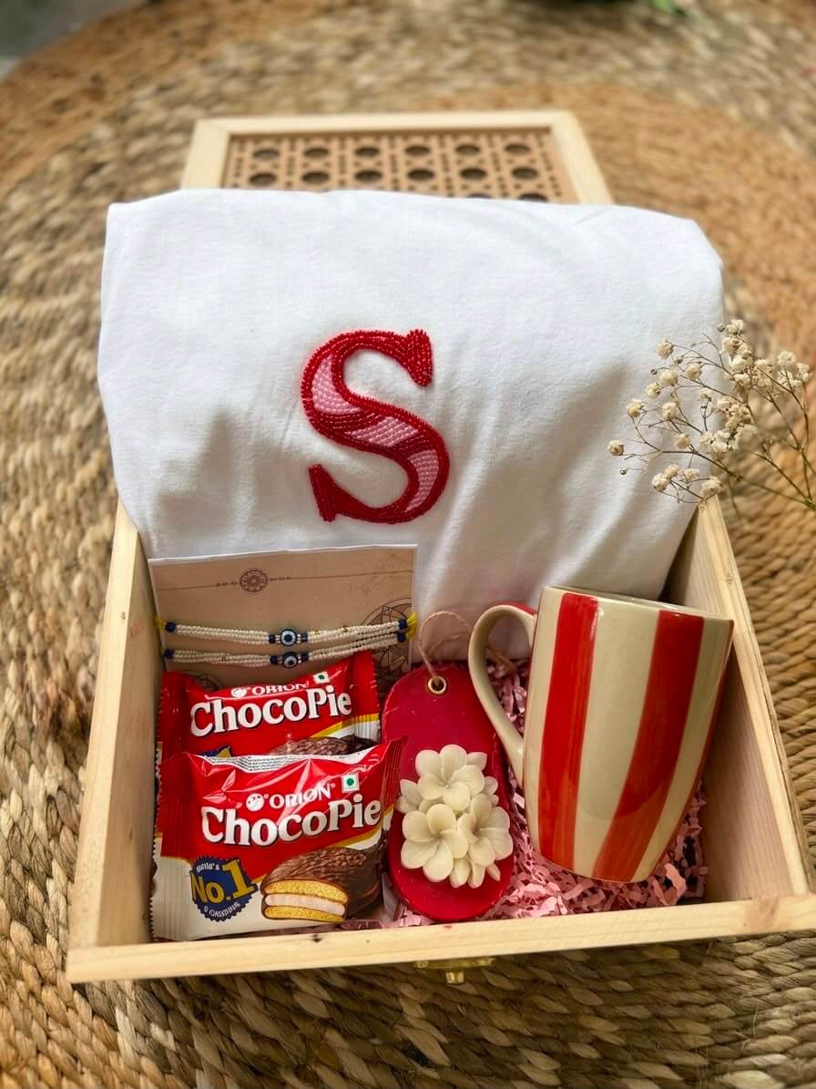 Gehru Cherish Box featuring a customized alphabet T-shirt, handcrafted Rakhi, red striped ceramic mug, floral sandal fragrance bar, Choco Pies, and premium wooden gift box.