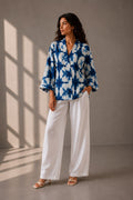 Woman wearing an indigo blue tie-dye cotton resort shirt with wide sleeves and white trousers.