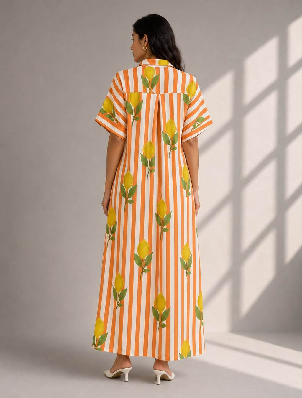 A woman viewed from the back wearing a long orange and white vertically striped dress with yellow floral prints, standing against a gray wall with soft window shadows.