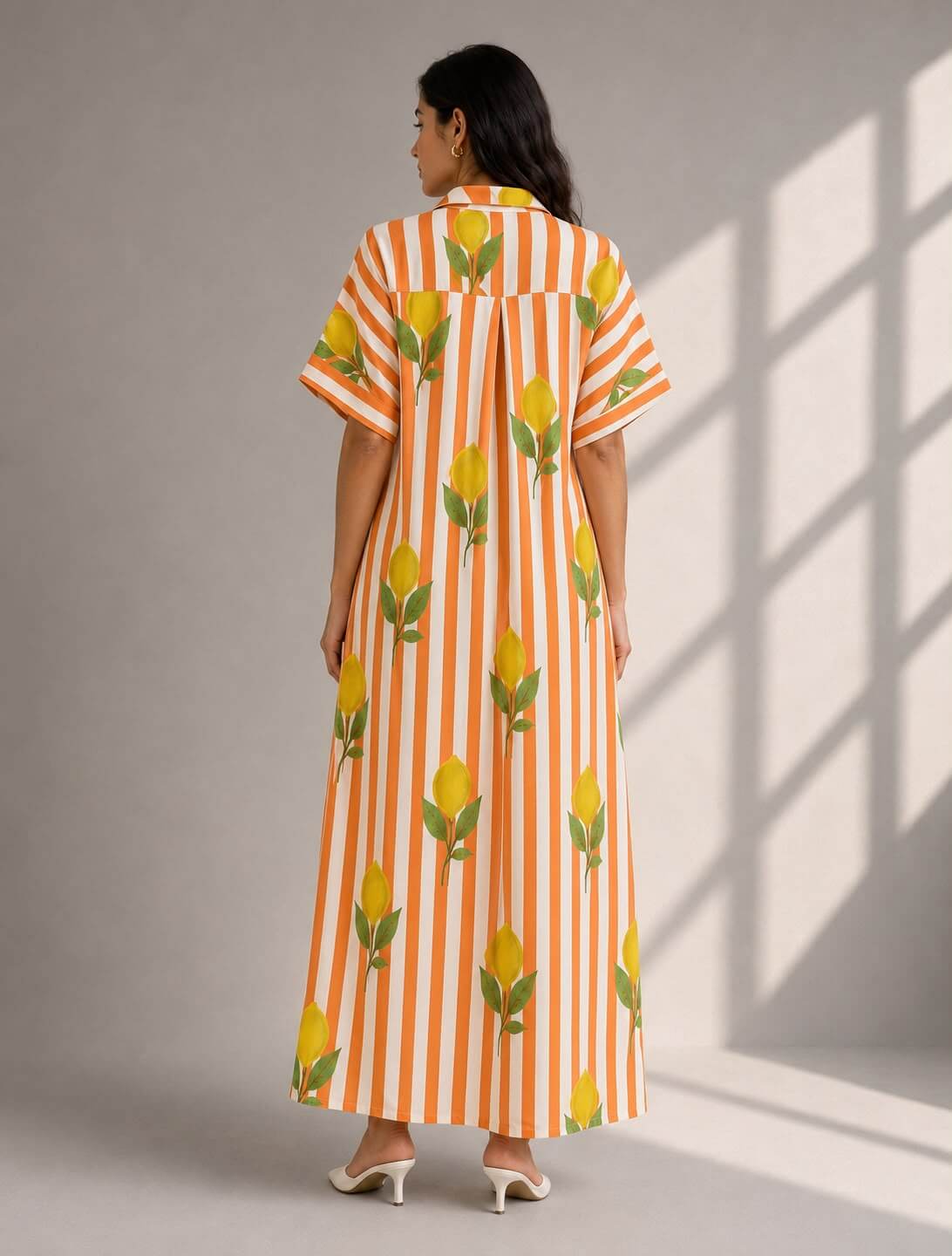 A woman viewed from the back wearing a long orange and white vertically striped dress with yellow floral prints, standing against a gray wall with soft window shadows.