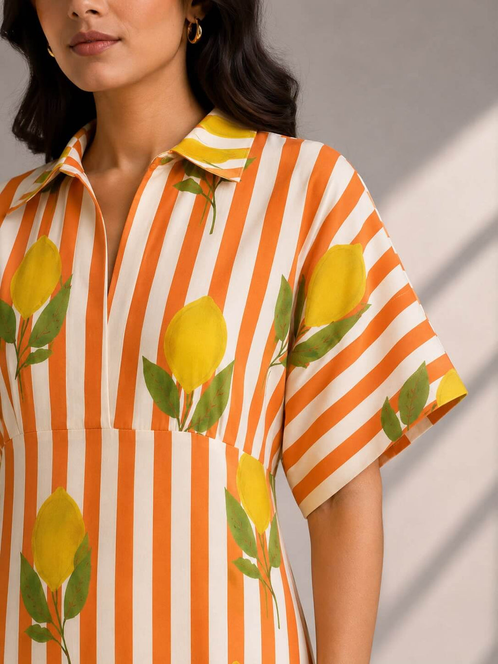 A close-up of a woman wearing a short-sleeve collared shirt dress with orange and white vertical stripes and a yellow lemon print.