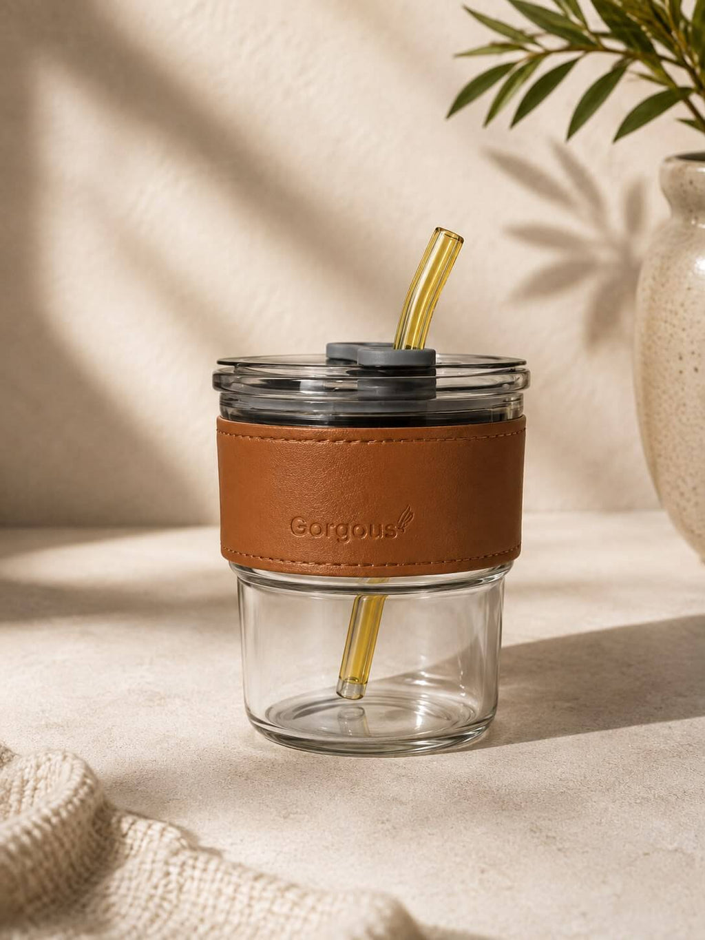 A clear glass travel tumbler with a brown leather sleeve embossed with the brand name 'Gorgous' and a gold-colored straw, shown on a textured beige surface with soft leaf shadows.