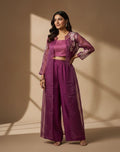 Woman in a purple traditional outfit standing against a beige wall with sunlight casting shadows.