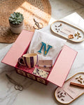 A styled gift box set on a marble surface, featuring a pink box containing a white embroidered monogram towel with a large letter 'N', a striped mug, a bar of soap, and a beaded bracelet. Decorative props including a small succulent, scissors, and gold-trimmed trays with spices are arranged around it.