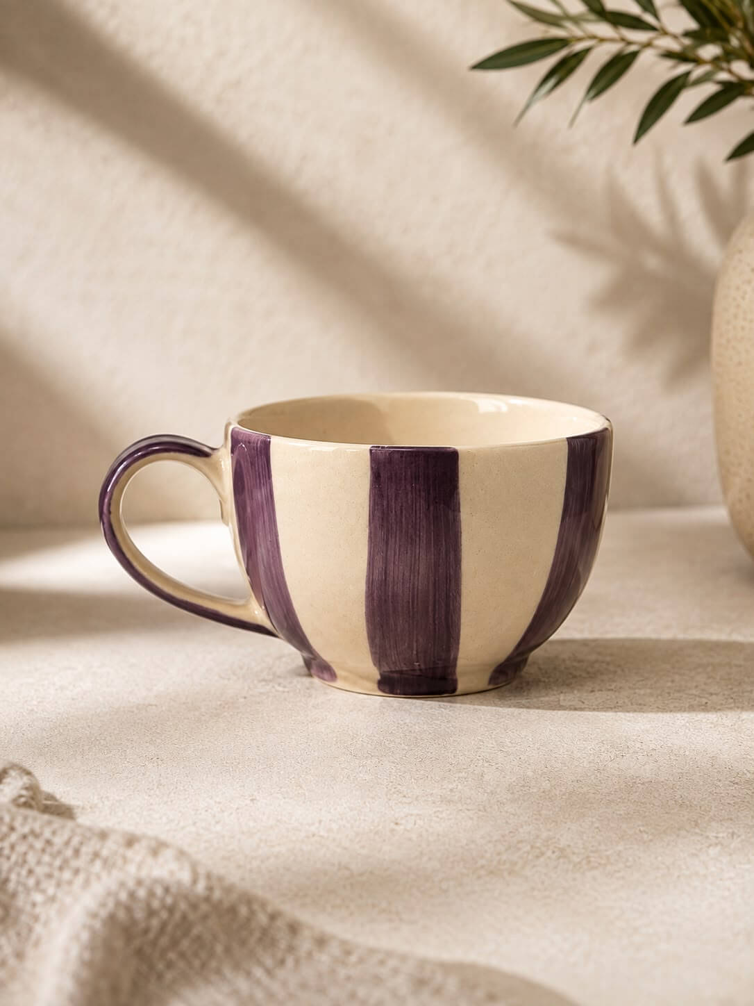 A ceramic coffee mug with a cream-colored interior and exterior featuring vertical hand-painted purple stripes, styled on a textured beige surface with soft shadows and a plant in the background.
