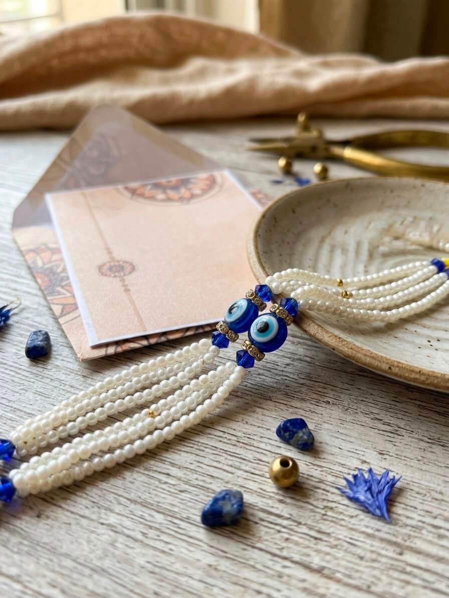 A bohemian-style multi-strand pearl rakhi with blue and white evil eye beads and dangling blue beads, styled on a rustic wooden table with a ceramic dish, gold-toned tools, and a card.