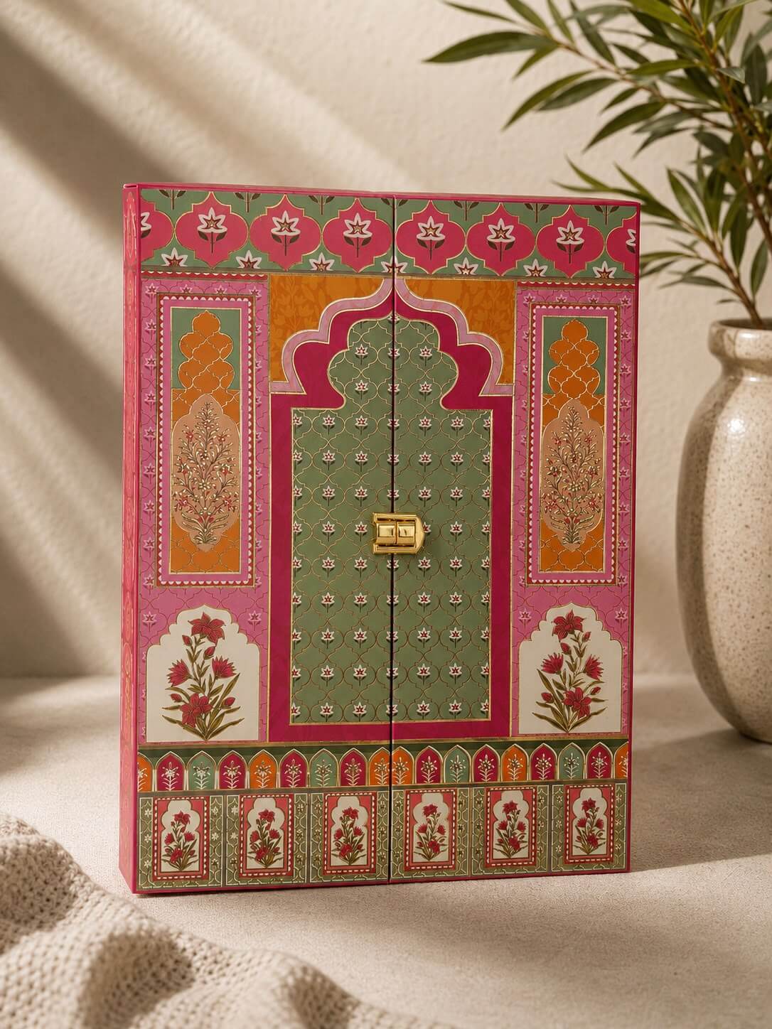 A decorative wooden storage box with intricate traditional Indian patterns in pink, green, orange, and gold, styled on a beige fabric surface next to a ceramic vase with green leaves.
