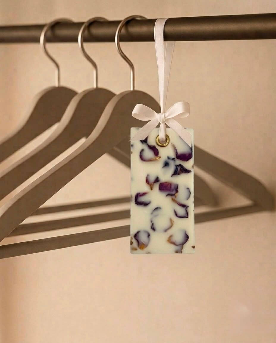 A rectangular white fragrance bar ornament with purple and brown botanical inclusions, tied with a white ribbon and hanging from a metal hanger on a clothing rack.