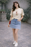 Woman wearing a beige t-shirt with a graphic design, denim skirt, and white sneakers in an outdoor setting.